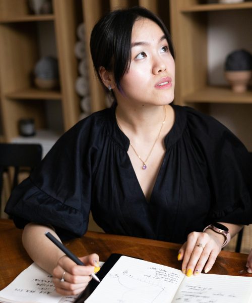 Asian woman entrepreneur writing notes at desk in office setting, focused and thoughtful.